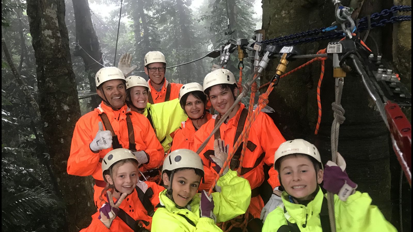 A smiling group in rain jackets and helmets on a misty zipline platform in the rainforest